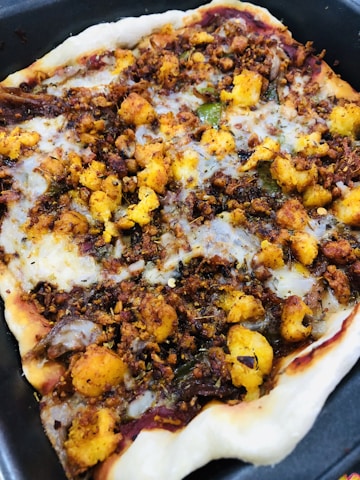 A close-up view of a homemade pizza topped with various ingredients such as fried cauliflower, spices, and bits of herbs on a thin crust. The edges appear slightly crisped, and the toppings are evenly spread across the surface, giving it a hearty and rustic appearance.