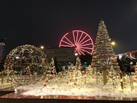 Festive decorations featuring illuminated structures resembling Christmas trees and a globe adorn a snowy landscape. In the background, a brightly lit Ferris wheel with a vivid red color stands out against the night sky. The scene is bustling with lights and colors, creating a warm and lively holiday atmosphere.
