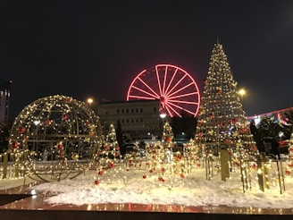 Festive decorations featuring illuminated structures resembling Christmas trees and a globe adorn a snowy landscape. In the background, a brightly lit Ferris wheel with a vivid red color stands out against the night sky. The scene is bustling with lights and colors, creating a warm and lively holiday atmosphere.