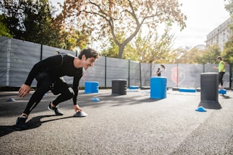 A man in athletic clothing is engaged in an outdoor workout or training session, bent forward while holding a marker or cone. The environment includes other sports equipment such as blue and gray barrels and more cones scattered around. In the background, additional people are seen participating, with trees and a building visible, suggesting a park or recreational area.
