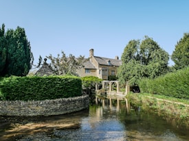 A picturesque stone cottage surrounded by lush greenery, including large trees and trimmed hedges, is reflected in a gently flowing stream. The clear blue sky adds to the tranquil atmosphere.