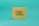 A small, portable, yellow speaker with a square shape featuring a grid pattern on its front surface. The logo 'JBL' is prominently displayed in large letters on the speaker's front. It is set against a solid teal background.