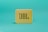 A small, portable, yellow speaker with a square shape featuring a grid pattern on its front surface. The logo 'JBL' is prominently displayed in large letters on the speaker's front. It is set against a solid teal background.