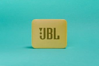 A small, portable, yellow speaker with a square shape featuring a grid pattern on its front surface. The logo 'JBL' is prominently displayed in large letters on the speaker's front. It is set against a solid teal background.