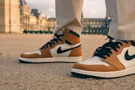 Close-up of a pair of stylish sneakers with a brown, black, and white color scheme worn by an individual standing on a gravel surface. In the background, an ornate historical building is visible, likely a museum or palace with classical architectural elements.