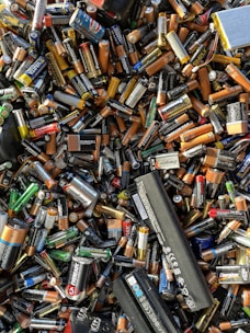 A large pile of assorted batteries, including AA, AAA, C, D, 9-volt, and rechargeable batteries from various brands like Duracell, Energizer, and Panasonic. The batteries are jumbled together in a chaotic manner with different colors and sizes visible.