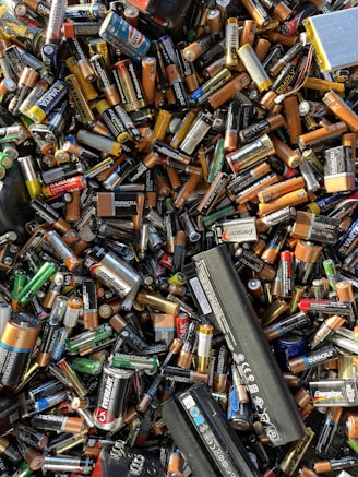 A large pile of assorted batteries, including AA, AAA, C, D, 9-volt, and rechargeable batteries from various brands like Duracell, Energizer, and Panasonic. The batteries are jumbled together in a chaotic manner with different colors and sizes visible.