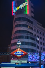 A nighttime cityscape featuring an illuminated metro sign labeled 'Callao' in front of a large building with a vibrant neon advertisement for 'Schweppes'. The background displays a classic architectural design with soft lighting and curved lines.