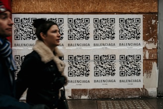 Multiple QR code posters are displayed on a wooden wall. Each poster contains a QR code with the word 'Balenciaga' printed beneath it. Two people are walking past the posters, slightly blurred, indicating motion.