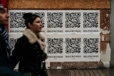 Multiple QR code posters are displayed on a wooden wall. Each poster contains a QR code with the word 'Balenciaga' printed beneath it. Two people are walking past the posters, slightly blurred, indicating motion.