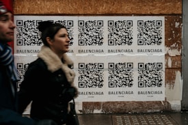 Multiple QR code posters are displayed on a wooden wall. Each poster contains a QR code with the word 'Balenciaga' printed beneath it. Two people are walking past the posters, slightly blurred, indicating motion.