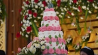 A multi-tiered white cake adorned with pink floral decorations and green leaves, topped with embellishments. Surrounding the cake are white and red flowers, along with green foliage, creating a festive backdrop.