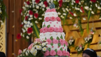 A multi-tiered white cake adorned with pink floral decorations and green leaves, topped with embellishments. Surrounding the cake are white and red flowers, along with green foliage, creating a festive backdrop.