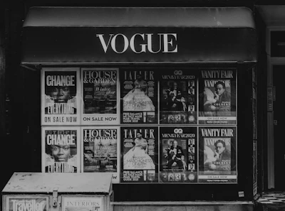Storefront with multiple magazine covers displayed in a window. The magazines include titles such as Vogue, House & Garden, GQ, and Vanity Fair. The covers feature various themes and personalities, with a focus on fashion, lifestyle, and culture.