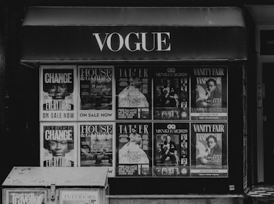 Storefront with multiple magazine covers displayed in a window. The magazines include titles such as Vogue, House & Garden, GQ, and Vanity Fair. The covers feature various themes and personalities, with a focus on fashion, lifestyle, and culture.