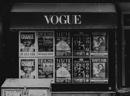 Storefront with multiple magazine covers displayed in a window. The magazines include titles such as Vogue, House & Garden, GQ, and Vanity Fair. The covers feature various themes and personalities, with a focus on fashion, lifestyle, and culture.