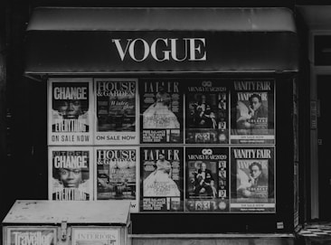 Storefront with multiple magazine covers displayed in a window. The magazines include titles such as Vogue, House & Garden, GQ, and Vanity Fair. The covers feature various themes and personalities, with a focus on fashion, lifestyle, and culture.