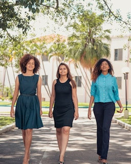 Three women confidently walk down a path lined with greenery and trees. They are dressed in smart and stylish outfits. Sunlight filters through the leaves, creating a natural and vibrant setting, with a building visible in the background.