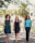 Three women confidently walk down a path lined with greenery and trees. They are dressed in smart and stylish outfits. Sunlight filters through the leaves, creating a natural and vibrant setting, with a building visible in the background.