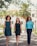 Three women confidently walk down a path lined with greenery and trees. They are dressed in smart and stylish outfits. Sunlight filters through the leaves, creating a natural and vibrant setting, with a building visible in the background.