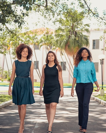 Three women confidently walk down a path lined with greenery and trees. They are dressed in smart and stylish outfits. Sunlight filters through the leaves, creating a natural and vibrant setting, with a building visible in the background.