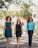 Three women confidently walk down a path lined with greenery and trees. They are dressed in smart and stylish outfits. Sunlight filters through the leaves, creating a natural and vibrant setting, with a building visible in the background.