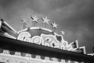 A classic carnival sign lit up with numerous light bulbs, featuring five large stars on top. The sign is designed in an ornate, vintage style with intricate patterns and swirling lines.