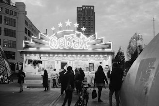 A black and white photograph of a nighttime urban scene with a brightly lit food stall decorated with stars and the word 'GEBÄK' on top. People are gathered around the stall, and a tall building is visible in the background. The scene includes elements of modern architecture and streetlights.