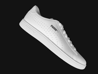 A sleek white sneaker with a minimalistic design features the Puma logo on the side. The shoe is positioned against a solid black background, highlighting its clean lines and smooth texture. Perforated details along the sides add a subtle touch of style.
