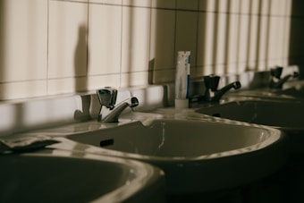 A row of white bathroom sinks with chrome faucets attached to a tiled wall. One of the sinks has a tube of toothpaste standing upright on its edge. The light coming through a window casts shadows and stripes on the wall and sinks, creating a somewhat somber mood.
