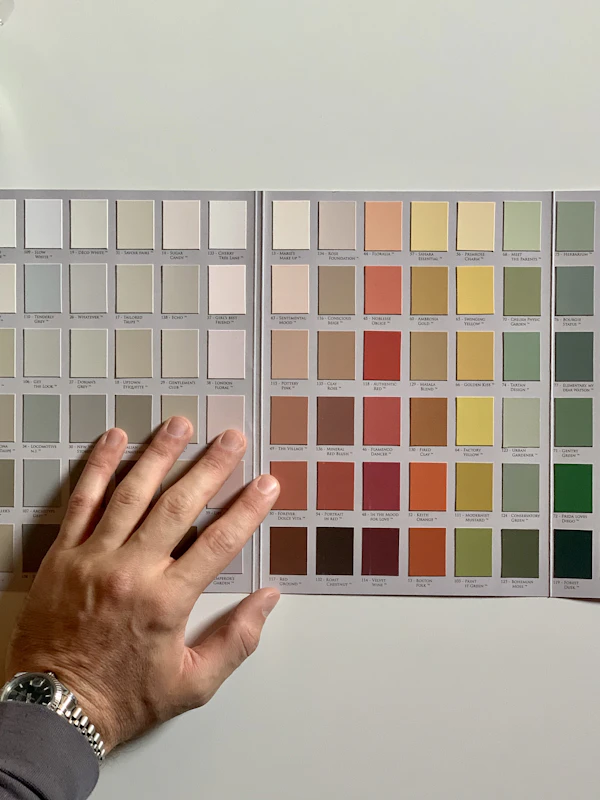 A person holding up a green paint sample card against a white interior wall.
