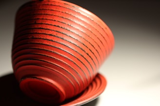 A beautifully crafted red ceramic cup with dark linear grooves encircling its surface, creating a striking pattern. The cup is set against a soft gradient background, casting a gentle shadow.