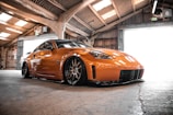 A sleek, modified orange sports car is parked inside an industrial-style garage. The car features custom rims and a low-slung body kit. The garage is illuminated by natural light coming through the translucent roof panels and wide-open garage doors.