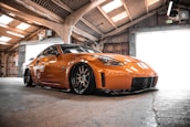 A sleek, modified orange sports car is parked inside an industrial-style garage. The car features custom rims and a low-slung body kit. The garage is illuminated by natural light coming through the translucent roof panels and wide-open garage doors.
