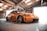 A sleek, modified orange sports car is parked inside an industrial-style garage. The car features custom rims and a low-slung body kit. The garage is illuminated by natural light coming through the translucent roof panels and wide-open garage doors.