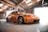 A sleek, modified orange sports car is parked inside an industrial-style garage. The car features custom rims and a low-slung body kit. The garage is illuminated by natural light coming through the translucent roof panels and wide-open garage doors.