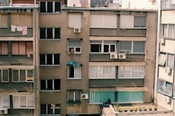 A residential apartment building facade with multiple windows, some of which have closed shutters. Clothes are hanging out to dry on a clothesline. Air conditioning units are installed in several windows, and a person is leaning out of one of the windows.