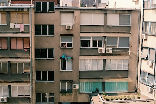 A residential apartment building facade with multiple windows, some of which have closed shutters. Clothes are hanging out to dry on a clothesline. Air conditioning units are installed in several windows, and a person is leaning out of one of the windows.