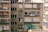 A residential apartment building facade with multiple windows, some of which have closed shutters. Clothes are hanging out to dry on a clothesline. Air conditioning units are installed in several windows, and a person is leaning out of one of the windows.