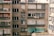 A residential apartment building facade with multiple windows, some of which have closed shutters. Clothes are hanging out to dry on a clothesline. Air conditioning units are installed in several windows, and a person is leaning out of one of the windows.