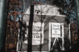 A wooden shed is situated in a forested area, with the trees displaying autumn colors. The shed bears a vintage-style sign that reads 'Jefferson Lakes Country Day Camp for Boys & Girls.' Adjacent to this, a door has a warning sign stating 'Private Property No Trespassing.' The atmosphere suggests a secluded, perhaps abandoned location with long shadows cast on the structure.