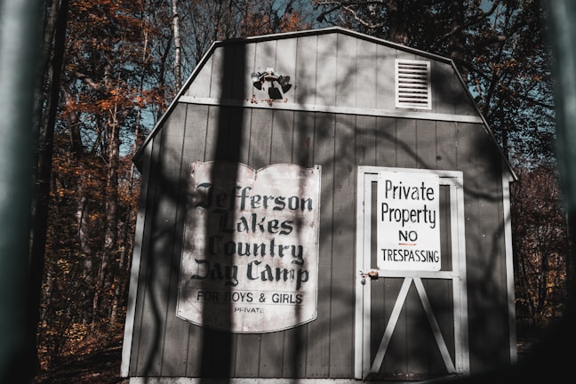 A wooden shed is situated in a forested area, with the trees displaying autumn colors. The shed bears a vintage-style sign that reads 'Jefferson Lakes Country Day Camp for Boys & Girls.' Adjacent to this, a door has a warning sign stating 'Private Property No Trespassing.' The atmosphere suggests a secluded, perhaps abandoned location with long shadows cast on the structure.