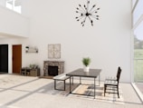 A modern dining room with minimalist decor features a sleek table and chairs on a large patterned rug. A potted plant sits centered on the table. The room is well-lit with large windows allowing natural light to enter. A stylish chandelier hangs from the ceiling. A stone fireplace, small console table, and decorative wall art complete the design.