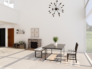 A modern dining room with minimalist decor features a sleek table and chairs on a large patterned rug. A potted plant sits centered on the table. The room is well-lit with large windows allowing natural light to enter. A stylish chandelier hangs from the ceiling. A stone fireplace, small console table, and decorative wall art complete the design.