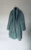 A long, green, fluffy coat hangs on a plain wall, suspended by a simple hanger. The coat has a cozy and textured appearance, suggesting warmth and comfort.