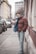 A young person stands casually on a city sidewalk, wearing a red puffer jacket and jeans. They lean slightly against a textured wall next to parked cars lining the street. The background features urban architecture, with buildings of varying heights and styles.