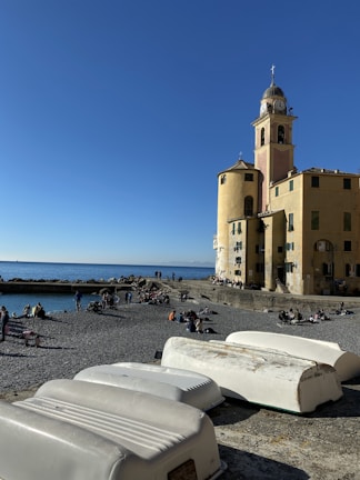 A coastal scene with a sandy and rocky beach featuring people relaxing and walking. In the foreground, several small white boats are overturned on the ground. A large, historic building with a clock tower stands prominently, overlooking the sea with a clear blue sky above.