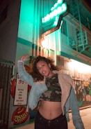 A confident person poses in front of a building with bright neon lights. The person is dressed in a denim jacket with a camo top underneath, and they stand with one arm raised. The building has a vintage feel, with a mural on the wall and an illuminated sign displaying 'Liquors' and 'Wine'.