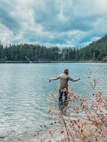 A person stands in the shallow waters of a lake, engaged in fly fishing with a rod. The individual is wearing outdoor clothing and a hat. Surrounding the lake are dense pine forests under a cloudy sky, and some dried foliage is visible in the foreground.