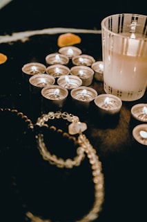 A collection of small tealight candles lit alongside a larger candle in a decorative holder creates a warm and inviting atmosphere. A beaded bracelet or string of beads is placed nearby, adding an element of personal or spiritual significance. The surface is dark, emphasizing the soft glow of the candlelight.
